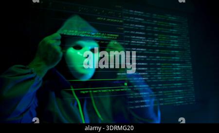 Anonymous hacker wearing a mask and hoodie interacting with digital data in a dark room Stock Photo