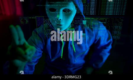Anonymous hacker wearing a mask and hoodie interacting with digital data in a dark room Stock Photo