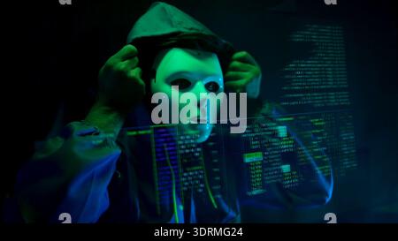 Anonymous hacker wearing a mask and hoodie interacting with digital data in a dark room Stock Photo