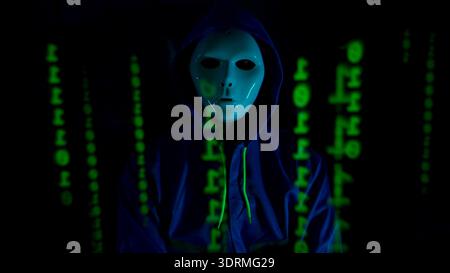 Anonymous hacker wearing a mask and hoodie interacting with digital data in a dark room Stock Photo