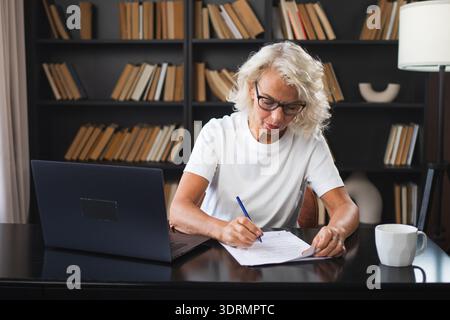 Middle aged senior woman using laptop computer writing notes at workplace. Focused mature old businesswoman work at office makin Stock Photo