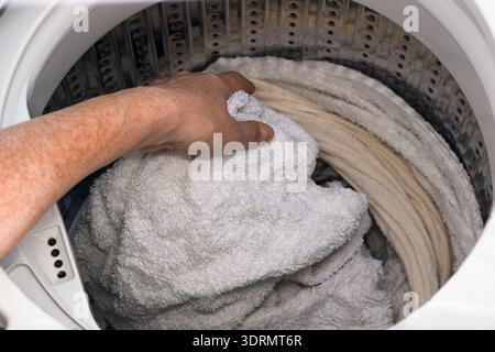 Closeup of woman hand taking wet laundry out of top load washing machine drum. Household chore concept showing fabric care, home cleaning routine, eve Stock Photo