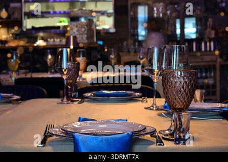 Elegant restaurant interior with neatly set tables, decorative colored goblets, wine glasses, white plates, and blue napkins. Warm lighting creates a Stock Photo
