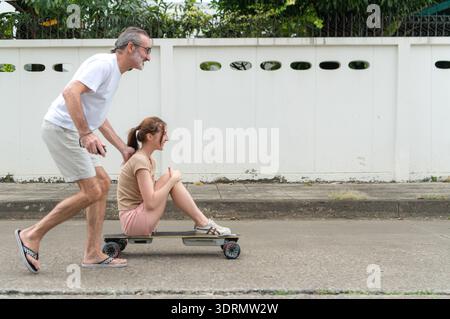 Elderly man playfully a young woman sitting on a skateboard on the street, showing fun, family bonding Stock Photo