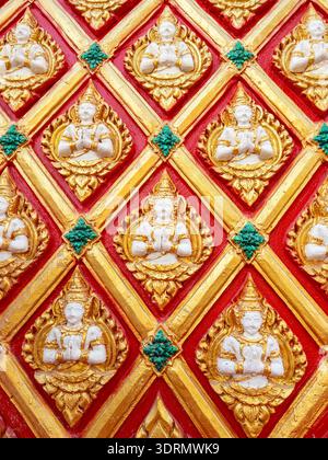 Decorative stucco relief featuring Thephanom angels in praying gesture on a red and gold traditional Thai temple wall. Intricate Buddhist art pattern Stock Photo