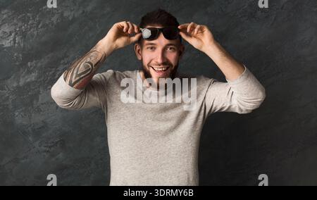 Man Holding Sunglasses and Smiling in Front of Dark Textured Wall Indoors With Casual Clothing Stock Photo