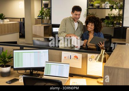 Collaborating diverse coworkers reviewing document behind office cubicle desk, with tablet. Collaboration, teamwork, modernoffic Stock Photo
