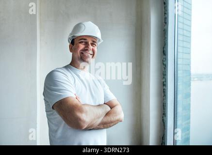 Smiling construction worker wearing a hard hat stands confidently with arms crossed, showcasing a bright and modern interior spa Stock Photo