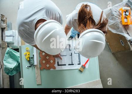 Two construction workers, a man and a woman, wearing white helmets, are reviewing blueprints on a table in a workshop, showcasin Stock Photo