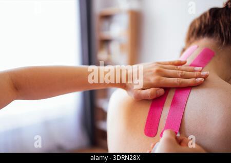 Female therapist applying pink kinesiology tape on the back of a client in a bright therapy room, showcasing therapeutic techniq Stock Photo