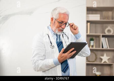 Mature senior male doctor in medical uniform using digital tablet in clinic, Therapist professional healthcare expert searching Stock Photo