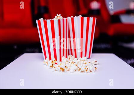 Two striped popcorn containers filled with fresh popcorn, placed on a white surface, with scattered popcorn and red cinema seats Stock Photo