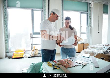 Two construction workers, a man and a woman, are discussing project plans in a bright, unfinished room filled with tools and mat Stock Photo