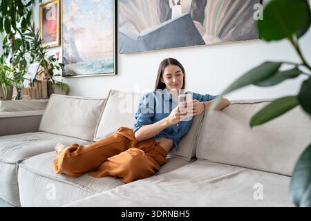 Happy relaxed woman artist sitting on couch at loft gallery apartment texting on smartphone online. Stock Photo