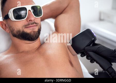 Male patient receiving laser hair removal treatment in a modern clinic, wearing protective eyewear, with a professional technici Stock Photo