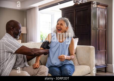 Diverse senior couple using blood pressure cuff on beige sofa in living room beside lamp, armoire. Health, wellness, senior, com Stock Photo