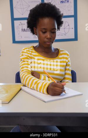 African American girl writing in notebook with pencil in classroom wearing yellow striped shirt. Student, education, classroom, Stock Photo