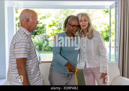 Diverse senior friends supporting woman using wooden cane in living room by sliding glass door. Senior, companionship, warm, inv Stock Photo