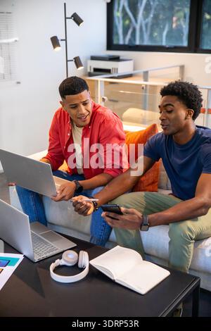 Diverse male friends collaborating using laptops, notebook, headphones, smartphone on sofa at home. Collaboration, innovation, m Stock Photo