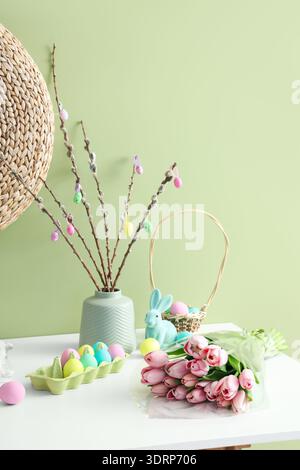 Vase with willow branches, Easter eggs and tulips on table near green wall Stock Photo