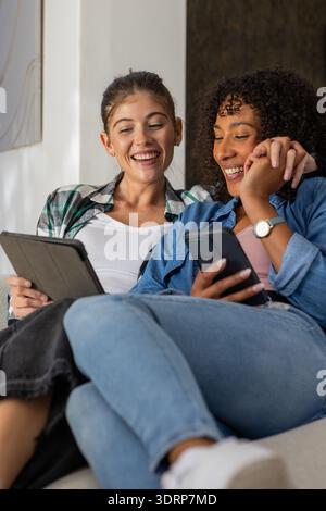 Diverse female friends reclining on light-colored couch in living room using tablet and smartphone. Casual, friendship, relaxati Stock Photo