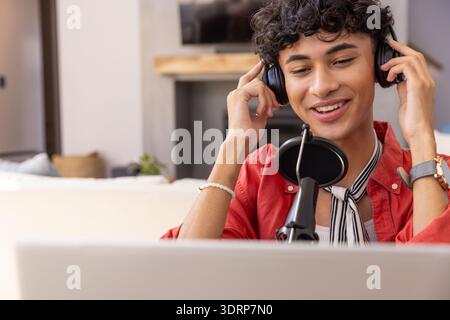 Man adjusting over-ear headphones while leaning toward cardioid microphone at desk in living room. Podcaster, content creator, h Stock Photo