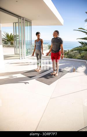 Diverse couple performing yoga on mats at modern balcony overlooking ocean with water bottles. Wellness, relaxation, serenity, t Stock Photo