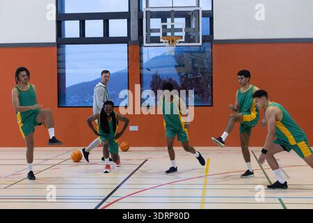 Basketball coach with five players in green jerseys performing lunges under hoop on gym court. Sportswear, teamwork, fitness, at Stock Photo