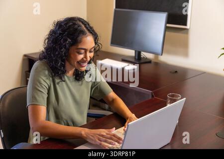 Indian woman typing on silver laptop at office desk with computer monitor plant bulletin board. Professional, workspace, modern, Stock Photo