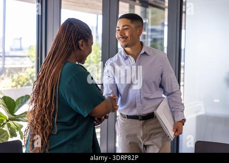 Diverse coworkers shaking hands while greeting in office near large windows with binder and plant. Professional, collaboration, Stock Photo