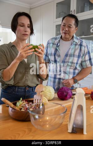 Diverse couple examining lettuce and slicing cabbage with mandoline slicer on kitchen countertop. Cooking, culinary, preparing, Stock Photo