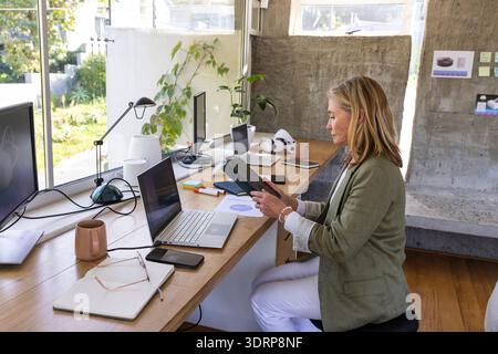Woman wearing light green blazer reading black notebook at wooden desk in home office with laptop. Workspace, professionalism, i Stock Photo
