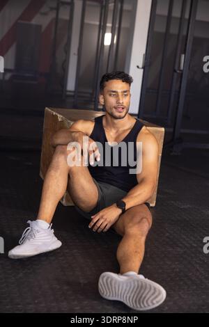 Asian man sitting on gym floor leaning against plyometric box wearing sportswear with squat racks. Fitness, strength, dedication Stock Photo