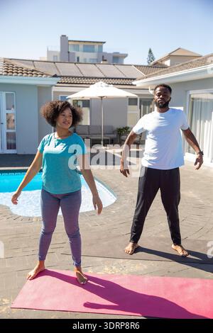 Diverse couple wearing sportswear practicing stretching exercises on yoga mats beside backyard pool. Fitness, wellness, outdoor, Stock Photo