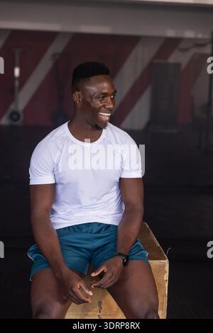 African American man sitting on wooden plyometric box at gym, holding smartwatch and smiling. Athlete, fitness, strength, traini Stock Photo