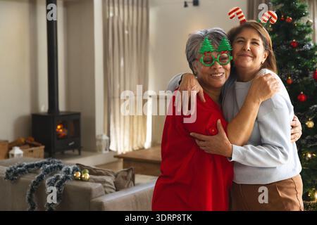 Diverse mother and daughter hugging in living room wearing festive glasses and candy cane headband. Family, celebration, cozy, s Stock Photo