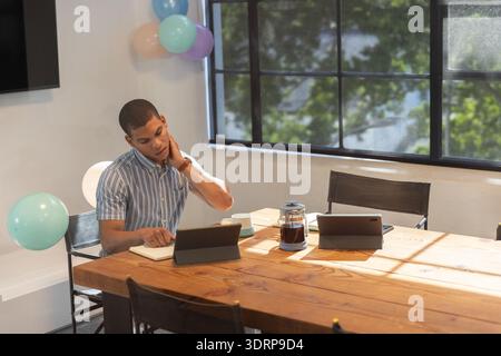 Man in mid-20s sitting at conference table at office using tablet, writing in notebook, copy space. Casual workspace, modern off Stock Photo