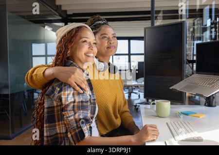 Diverse female coworkers celebrating while hugging at office desk with monitors and coffee mug. Collaboration, teamwork, diversi Stock Photo