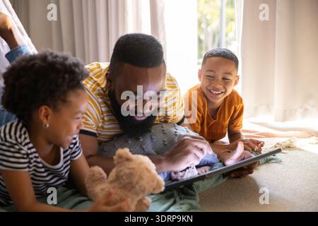 Diverse family lying on rug in living room using tablet computer with pillows, stuffed teddy bear. Family, relaxation, bonding, Stock Photo