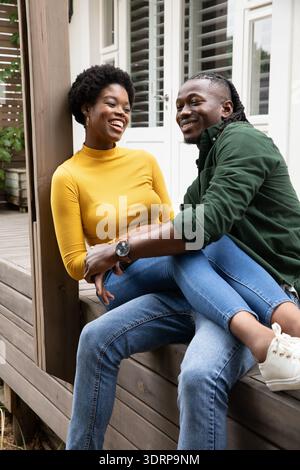 African American couple sitting close and smiling on porch bench with shuttered windows. Romantic, leisure, lifestyle, cozy, aut Stock Photo