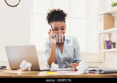 Young Woman Speaks on Phone While Taking Notes at Desk in Bright Office Stock Photo