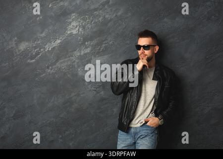 Man Poses in Leather Jacket Against Gray Wall With Sunglasses on During Casual Indoor Session Stock Photo