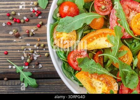 Fresh salad of red, pink and yellow tomatoes with arugula and spices in a white plate on a dark wooden table. Selective focus Stock Photo