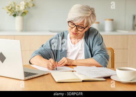 Middle aged senior woman using laptop computer writing notes at home. Focused mature old businesswoman work in home office makin Stock Photo