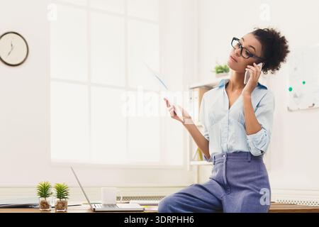 Young Woman Speaks on the Phone While Holding Paper and Relaxing in an Office Setting Stock Photo