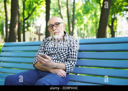 Elderly Man Sits on a Bench in a Park Checking His Phone on a Sunny Day Stock Photo