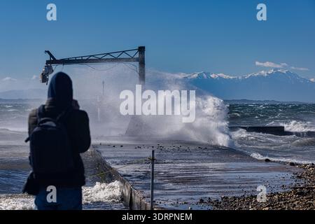 A person stands on a coastal pier as powerful waves crash against the structure, sending water into the air. A metal crane rises above the scene, whil Stock Photo