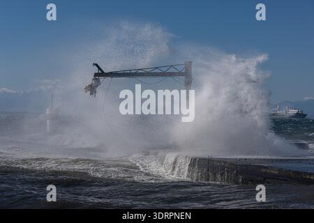 Powerful waves strike the concrete pier, splashing around a metal crane structure as the snow‑covered peak of Mount Olympus rises in the background. T Stock Photo