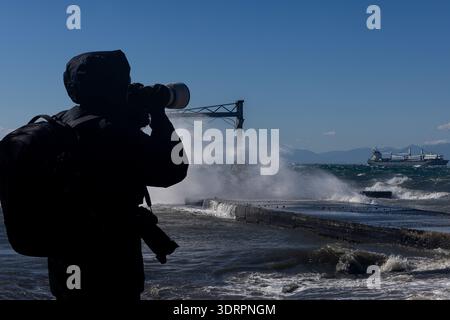 A photographer with a telephoto lens stands near a stormy pier as powerful waves crash against the structure. A cargo ship sails through rough waters Stock Photo