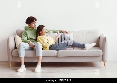 Affectionate young Asian couple using smartphone, relaxing on couch together at home, free space Stock Photo